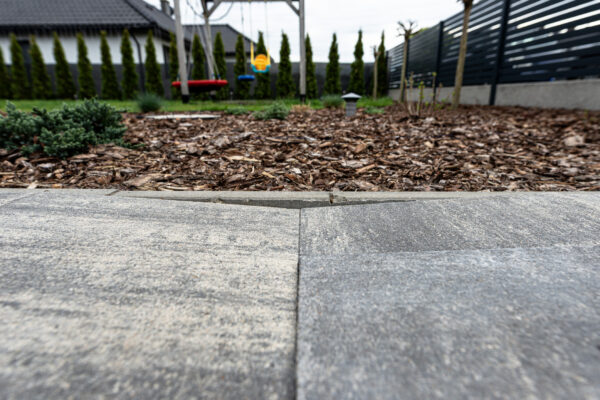 Close-up view of gray concrete pavers in the foreground, with a mulch-covered garden and blurred backyard playground equipment in the background.
